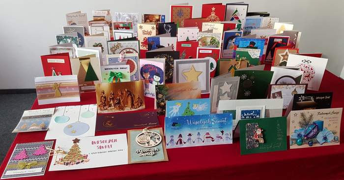 Pokaż zdjęcie: Na czerwonym płótnie rozłożone zostały różnorodne kartki z życzeniami świątecznymi zawierające akcenty bożonarodzeniowe, z czego cześć z nich została wykonana przez osoby niepełnosprawne w ramach rękodzieła artystycznego 