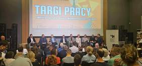 Pokaż zdjęcie: Na scenie siedzą różne osoby uczestniczące w panelu dyskusyjnym. Na widowni znajduje się tłum ludzi. Na ekranie napisana jest nazwa wydarzenia: Targi Pracy i Możliwości dla Osób z Niepełnosprawnościami. 