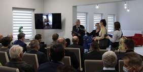 Pokaż zdjęcie: na zdjęciu grupa osób siedzi i ogląda prezentację powstawania obiektu ŚDS, po drugiej stronie stoi Dyrektor Oddziału Łódzkiego PFRON , odczytuje list z podziękowaniami od Prezesa Zarządu PFRON