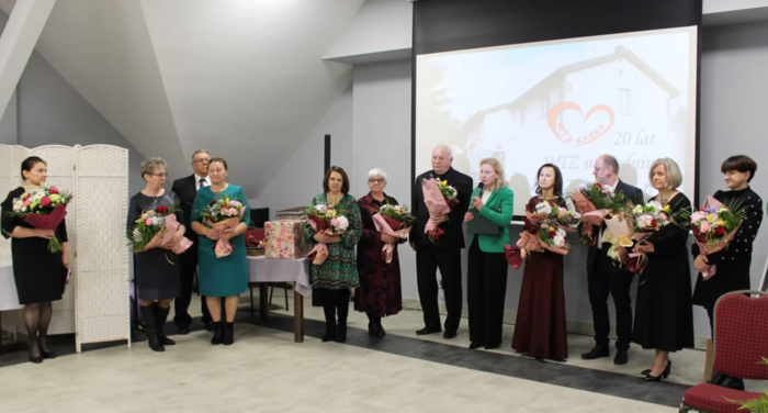 Pokaż zdjęcie: W sali po okręgu stoi duża grupa osób odświętnie ubranych, większość osób trzyma w ręku kwiaty. Mikrofon trzyma kobieta blondynka w zielonej marynarce.