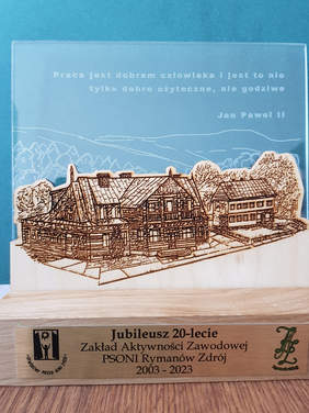 Jubileuszowa statuetka na szkle widok budynków ZAZ, na dole napis jubileusz 20 lecia ZAZ, logo PSONI, logo ZAZ u góry napis: praca jest dobrem człowieka i jest to nie tylko dobro użyteczne, ale godziwe Jan Paweł II 