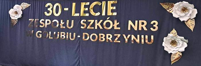 Na granatowym tle widnieje napis: 30-lecie Zespołu Szkół nr 3 w Golubiu-Dobrzyniu. Wokół wiszą duże białe kwiaty ze złotymi liśćmi