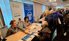 Pokaż zdjęcie: Dwie kobiety siedzą przy stole na którym leżą informatory i materiały promocyjne. Kobiety udzielają porad dwóm kobietom. Pierwsza kobieta siedzi druga stoi, za nimi widać stojący tłum ludzi. 