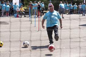 Pokaż zdjęcie: Uczestnik Spartakiady kopie piłkę podczas konkurencji piłkarskiej na torze przeszkód.  W tle inni uczestnicy i kibice obserwują zawody.