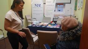 Pokaż zdjęcie: W pokoju znajdują się dwie kobiety: starsza pani siedząca na krześle i stojąca obok młoda dziewczyna. Starsza kobieta trzyma w dłoni informator dotyczący zdrowia. W tle widać stolik z ulotkami i materiałami reklamowymi. 