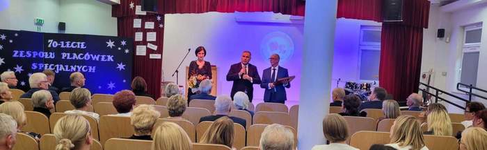 Pokaż zdjęcie: Na scenie stoi mężczyzna ubrany w garnitur krawat i białą koszulę trzyma mikrofon i mówi. Po jego prawej mężczyzna również na galowo ubrany trzyma w ręku grawer. Po lewej stoi kobieta, która trzyma kwiaty.