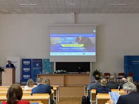 Pokaż zdjęcie: Sala wykładowa; uczestnicy siedzą w ławkach, przed nimi mównica i ekran z prezentacją „Przyjazne miejsce pracy”. Obok stoją roll-upy organizatorów, a prelegent przemawia.