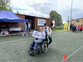 Pokaż zdjęcie: Na wózku inwalidzkim siedzi uśmiechnięta dziewczyna w okularach, w ręku trzyma białą kartkę z drukowanym napisem WTZ Lębork. Za nią stoi dziewczyna ubrana w czapeczkę z daszkiem również uśmiecha się do zdjęcia. W tle niebieski namiot, pod k