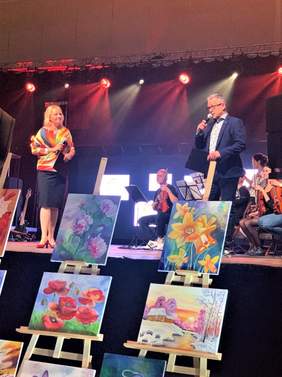 Pokaż zdjęcie: Na scenie stoja prowadzący - kobieta i mężczyzna. Pod sceną na sztalugach ustawione są obrazy.