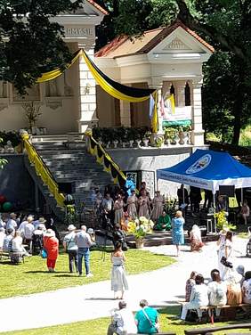 Pokaż zdjęcie: Zdjęcie z większej odległości obejmujące duży budynek oraz zbudowaną przed nim scenę, na scenie trwa występ artystyczny.