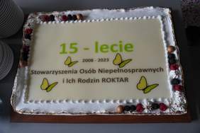 Pokaż zdjęcie: Na stole leży tort udekorowany malinami i jeżynami. Na torcie widoczne są również następujące sformułowania: „15-lecie”; „2008-2023” oraz „Stowarzyszenia Osób Niepełnosprawnych i Ich Rodzin ROKTAR”, jak również żółte motyle. Po lewej stronie możemy dostrzec dwa komplety talerzy.