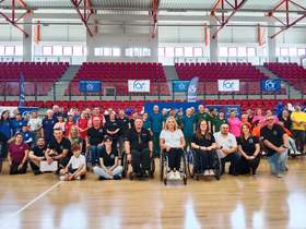 W hali sportowej stoi tłum ludzi. Wszyscy są ustawieni w rzędach i pozują do zdjęcia. W tle widać trybuny. 