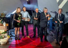 Pokaż zdjęcie: Na scenie stoi czterech mężczyzn i jedna kobieta. Odbywa się wręczanie nagród i dyplomów. Wszyscy są elegancko ubrani. 