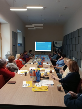 Pokaż zdjęcie: Grupa uczestników siedzi przy dużym stole w sali konferencyjnej, na którym leżą materiały informacyjne, napoje i przekąski. Na ścianie wyświetlana jest prezentacja, a w tle widnieje baner organizatora wydarzenia.