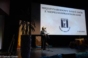 Pokaż zdjęcie: Zdjęcie ze sceny. Na pierwszym planie po prawej stronie stoi kobieta ubrana w czarny żakiet i spodnie, przemawiająca do mikrofonu, trzymając kartki. Za nią na dużym ekranie widnieje napis "MIĘDZYNARODOWY DZIEŃ OSÓB Z NIEPEŁNOSPRAWNOŚCIAMI" oraz logo i data "3 XII 2025". Po lewej stronie ekranu stoi duża donica z rośliną.