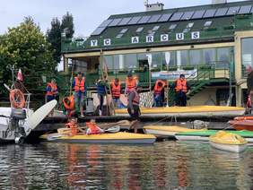 Pokaż zdjęcie: Uczniowie podczas szkolenia żeglarskiego dwie kobiety w kajaku, mężczyźni stoją na pomoście w dłoniach trzymają wiosła.