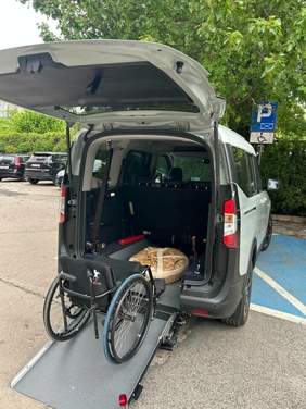 Pokaż zdjęcie: Na parkingu stoi samochód przystosowany do przewozu osób z niepełnosprawnościami. Tylna klapa pojazdu jest otwarta. Na rampie samochodowej stoi ręczny wózek inwalidzki. 