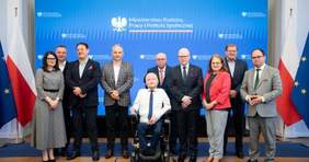 Pokaż zdjęcie: Przed niebieskim bilbordem jest dziesięć osób: dwie panie i ośmiu panów. Dziewięć osób stoi, a jeden mężczyzna siedzi na elektrycznym wózku inwalidzkim. Na bilbordzie jest napisane: Ministerstwo Rodziny, Pracy i Polityki Społecznej. 