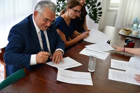 Pokaż zdjęcie: Przy stole w kolorze ciemnego drewna, szpakowaty mężczyzna w ciemnym garniturze podpisuje umowę, obok siedzi kobieta w ciemnej bluzce, przegląda dokumenty