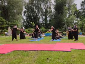 Pokaż zdjęcie: aktorzy ubrani na czarno stoją za różową matą rozłożoną na trawie przeciągają między sobą liny