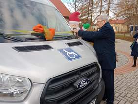 Pokaż zdjęcie: Dyrektor Łódzkiego Oddziału PFRON przecina wstęgę, którą jest otoczony samochód. W tle widać nadmuchanego Świętego Mikołaja i osobę robiącą zdjęcie. 
