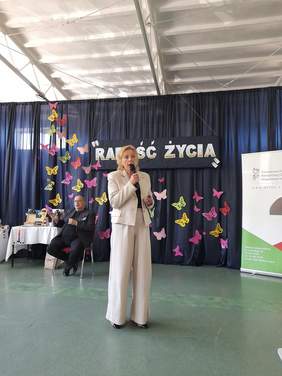 Pokaż zdjęcie: Podczas konferencji kobieta w jasnym garniturze, prowadzi wykład na temat sprzętów dostępnych w Ośrodku Wsparcia i Testów w Świdniku.