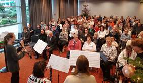 Pokaż zdjęcie: Grupa około 100 kobiet i mężczyzn, z czego część z tych osób porusza się na wózku, siedzi w sali konferencyjnej i wysłuchuje koncertu zespołu klarnetowego podczas finału IV edycji Konkursu Oddziału Wielkopolskiego PFRON na kartkę bożonarodzeniową.