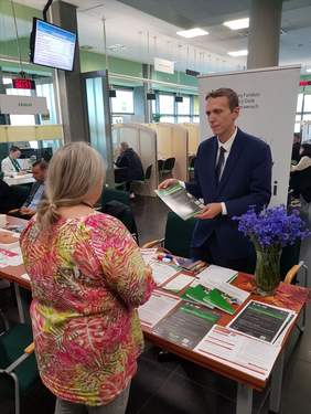 Pokaż zdjęcie: Mężczyzna stoi za stolikiem, na tle baneru PFRON i udziela informacji kobiecie, która przyszła na Dzień Seniora. Na stoliku ustawiony jest bukiet z chabrami oraz ułożone są materiały promujące programy PFRON. Stół udekorowany jest kolorowymi liśćmi. 