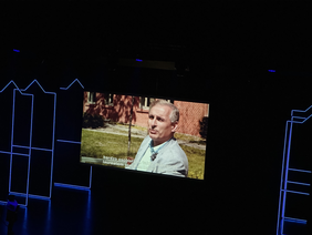 Pokaż zdjęcie: Potężny telebim na ciemnej scenie z delikatnym doświetleniem. Na ekranie mężczyzna w szarej marynarce  i jasne koszuli na tle parku i budynku z czerwonej cegły siedzi bokiem do kamery.