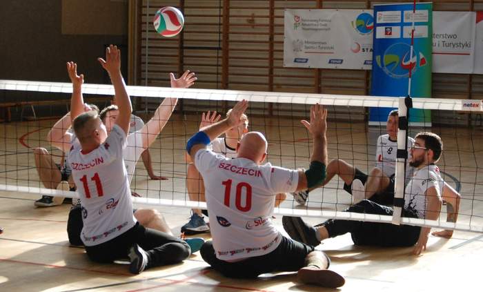 Pokaż zdjęcie: Siędzący na boisku zawodnicy w strojach sportowych przedzieleni nisko zawieszoną siatką, w powietrzu znajduje się rzucona piłka do siatkówki