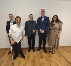 Pokaż zdjęcie: Pięć osób pozuje do wspólnego zdjęcia na tle białej ściany.