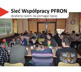 Sala wypełniona dorosłymi uczestnikami siedzącymi przy stołach, zwróconymi w stronę prowadzącego. W lewym górnym rogu widnieje hasło „Sieć Współpracy PFRON – działamy razem, by pomagać lepiej”.