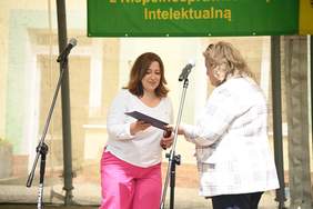 Pokaż zdjęcie: Na scenie stoją dwie kobiety, jedna drugiej wręcza list gratulacyjny.