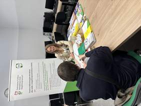 Pokaż zdjęcie: Konsultacja przy stoisku informacyjnym. Kobieta w jasnym żakiecie rozmawia z mężczyzną siedzącym tyłem. Na stole leżą materiały informacyjne i koszyk z cukierkami. W tle stoi roll-up z napisem: „PFRON – Centra informacyjno-doradcze dla osób z niepełnosprawnością”.