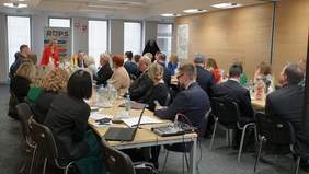 Pokaż zdjęcie: Dwa stoły ustawione w podkowę, przy których trwa posiedzenie, przy stole siedzi duża grupa osób.  Z tyłu roll-up Regionalnego Ośrodka Polityki Społecznej. Na zdjęciu stoi i zabiera głos dyrektor ROPS Pani Małgorzata Romanko ubrana w czerwona marynarkę.