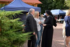 Pokaż zdjęcie: Na dworze przy zielonym krzaku stoją trzy osoby, zakonnica, kobieta i mężczyzna, rozmawiają ze sobą.