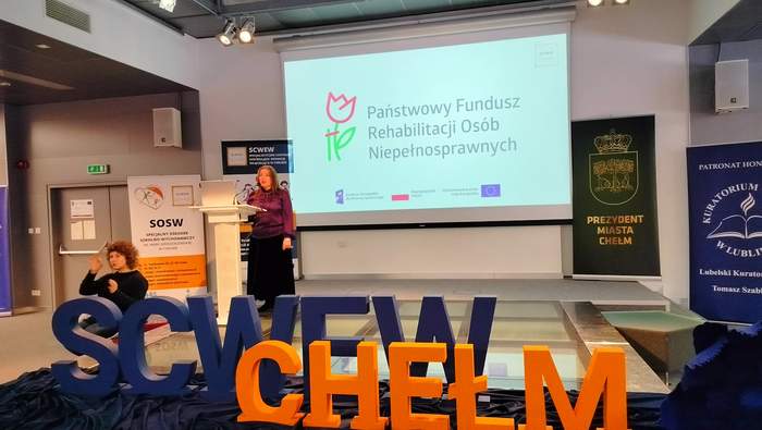 Pokaż zdjęcie: Prelegentka stojąca przy mównicy podczas konferencji SCWEW w Chełmie. Na ekranie widoczne logo Państwowego Funduszu Rehabilitacji Osób Niepełnosprawnych. Na scenie duże litery SCWEW Chełm i banery organizatorów.