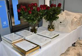 Pokaż zdjęcie: Na stole przykrytym białym obrusem leżą dyplomy oprawione w białe ramki formatu A4, na stole stoi wazon z czerwonymi różami oraz siatki lniane z napisem pralnia TARA
