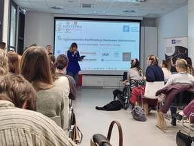 Pokaż zdjęcie: Konferencja naukowo-szkoleniowa – prelegentka stoi na tle ekranu z napisem „V Ogólnopolska Konferencja Naukowo-Szkoleniowa – Dostępna Ginekologia: Otwartość w dialogu”, przed nią publiczność, w tym osoba na wózku z psem asystującym.  