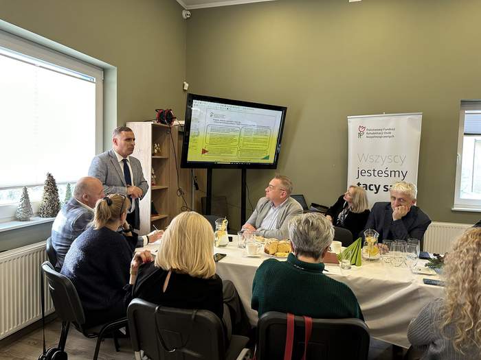 Pokaż zdjęcie: Mężczyzna w szarej marynarce i krawacie mówi do osób siedzących przy długim stole. Obok duży TV w prezentacją i baner z logo PFRON.