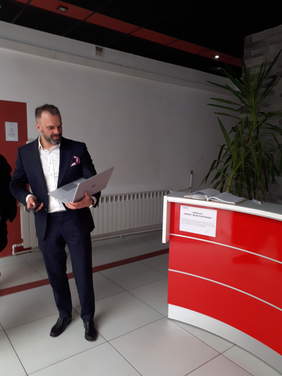 Pokaż zdjęcie: Hol wejściowy z ladą recepcji, na pierwszym planie ekspert  BHP trzyma notebooka i dalmierz laserowy