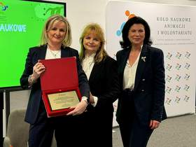 Trzy kobiety, ubrane elegancko, uśmiechnięte. Pozują do zdjęcia z grawerowanym dyplomem pamiątkowym w czerwonym etui.