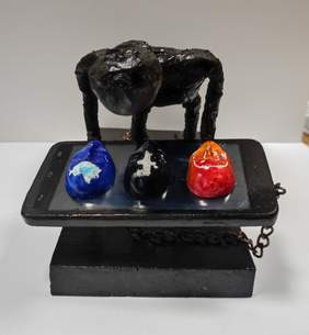 Pokaż zdjęcie: Zdjęcie rzeźby w kolorze czarnym przedstawiająca człowieka klęczącego nad telefonem i przykutego do niego łańcuchami. Na telefonie są trzy twarze z logo facebook, instagram i twitter