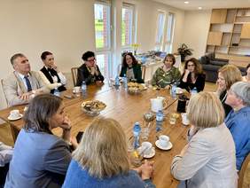 Pokaż zdjęcie: Liczna grupa osób siedzi przy dużym drewnianym stole. Na stole kawa, herbata, woda i ciastka. Spotkanie odbywa się w przestronnym, jasnym pomieszczeniu z dużymi oknami i regałem w tle.