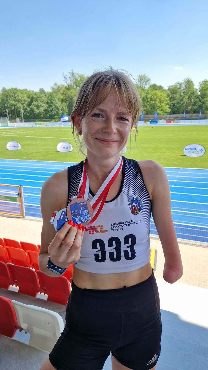 Pokaż zdjęcie: Młoda dziewczyn w sportowym stroju pozuje do zdjęcia na tle stadionu lekkoatletycznego trzymając w prawej ręce medal 