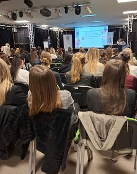 Pokaż zdjęcie: Sala konferencyjna. Na krzesłach ustawionych w rzędach siedzą uczestnicy spotkania zwróceni twarzami w kierunku sceny. W głębi sali znajduje się scena wraz z ekranem.