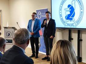Dwaj mężczyźni stoją obok siebie na tle niebieskiego baneru „Powiat Policki”. Mężczyzna po prawej, młodszy, ubrany w ciemny garnitur i białą koszulę, mówi do mikrofonu. Mężczyzna po lewej, starszy, w okularach i jasnoniebieskiej marynarce, słucha go z zaplecionymi dłońmi. Obok nich po prawej stronie znajduje się duży ekran wyświetlający okrągłe, niebiesko-białe logo z napisem w otoku: „SOSW NR 1 DLA DZIECI NIEPEŁNOSPRAWNYCH RUCHOWO • POLICE”. Na pierwszym planie widoczne są nieostre sylwetki i tyły głów siedzących tyłem do obiektywu słuchaczy.