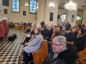 Pokaż zdjęcie: W kościele trwa koncert kolęd. Po lewej stronie stoją artyści. Po prawej stronie siedzą w ławkach ludzie, którzy słuchają wykonania kolęd i pastorałek. 