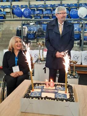 Pokaż zdjęcie: Obok siebie są kobieta i mężczyzna. Pan stoi, a pani siedzi na ręcznym wózku inwalidzkim. Przed nimi znajduje się na stoliku tort z zapalonymi racami. W tle widać halę sportową. 