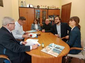 Pokaż zdjęcie: Przy owalnym stole siedzą dwie kobiety i czterech mężczyzn. Rozmawiają ze sobą i podpisują umowę. Na stole leżą różne dokumenty i pieczątki. 